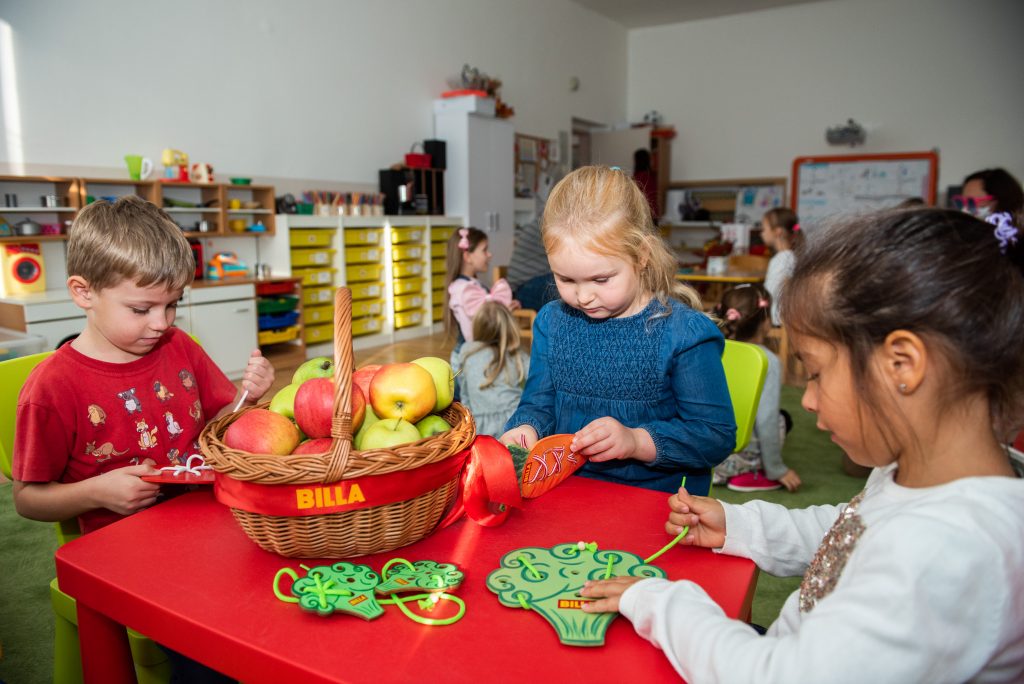 Deti sú za stolom, pred nimi košík s ovocím a v rukách majú hračky v tvare ovocia a zeleniny.
