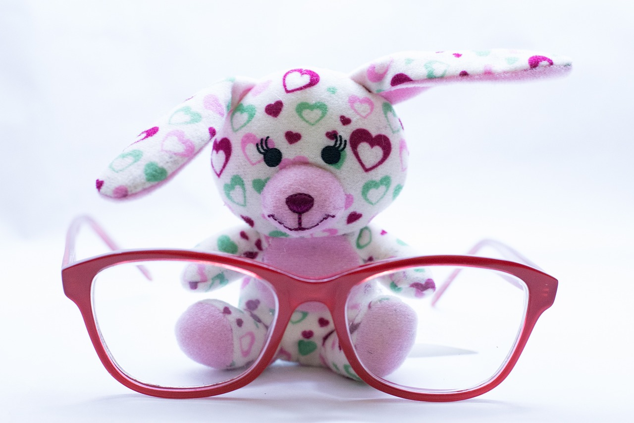 Na obrázku je biela plyšová hračka zajačika, na ktorom sú farebné srdiečka a pred ním sú detské okuliarky červenej farby .