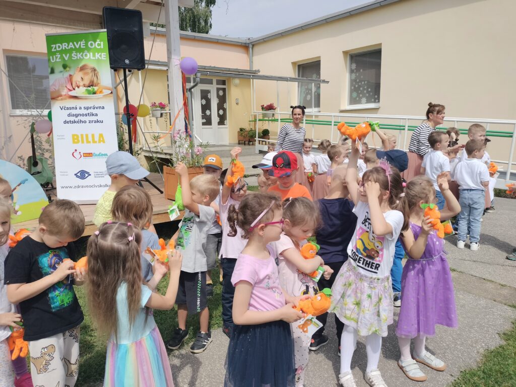 Na obrázku sú deti s pani učiteľkami, v rukách majú plyšové mrkvičky a hrášky. Na pódiu vidno rollup s textom Zdravé oči už v škôlke, Včasná a správna diagnostika detského zraku, BILLA, BILLA ľuďom, Únia nevidiacich a slabozrakých Slovenska + logá a web www.zdraveocivskolke.sk